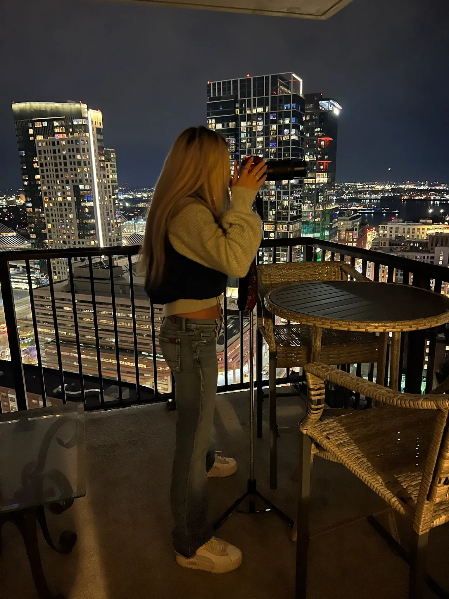 Gabriella on a high-rise balcony at night, looking through a telescope over the city lights