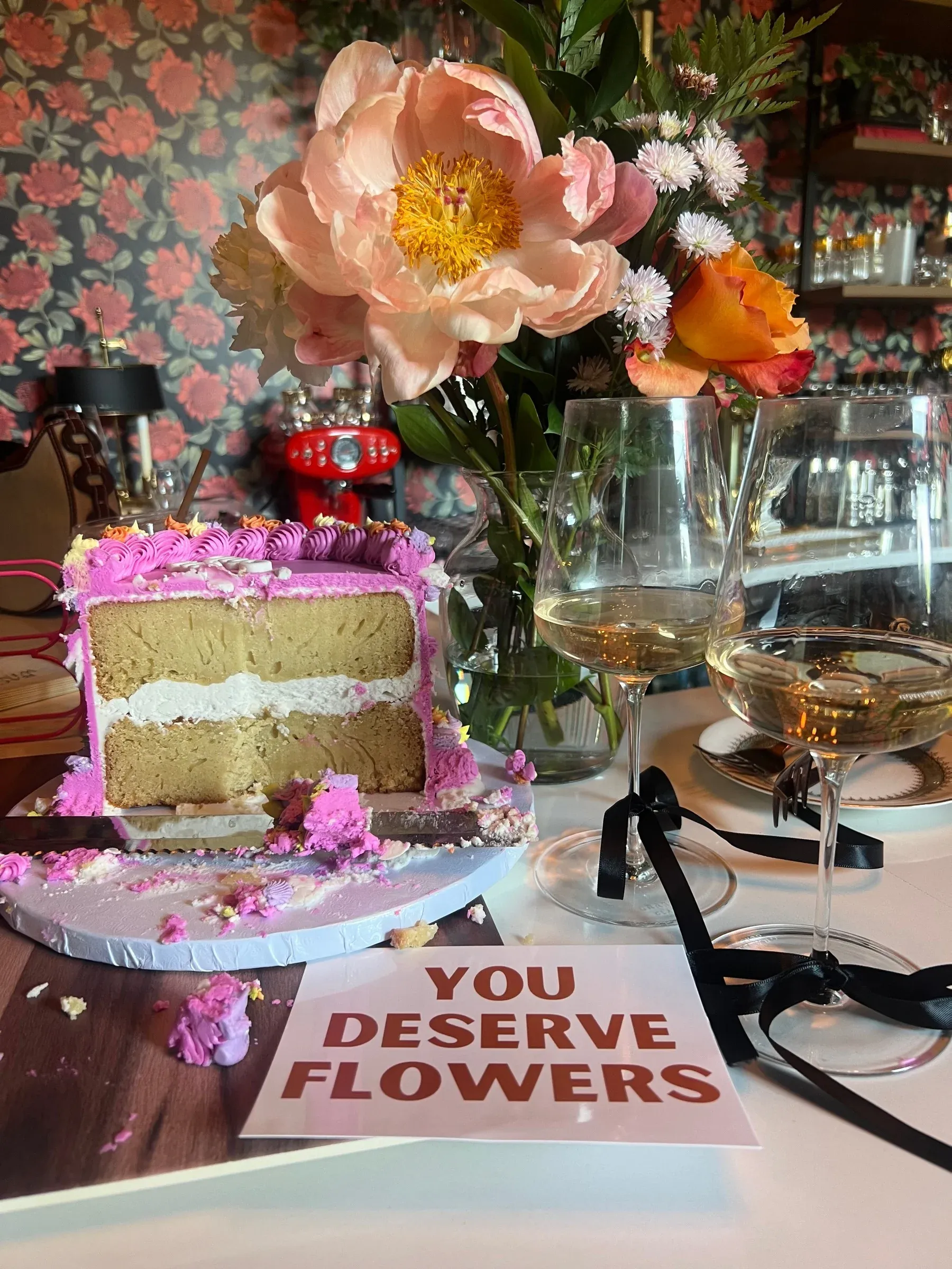 Still life of peonies, a layered cake and a “You deserve flowers” card on a table