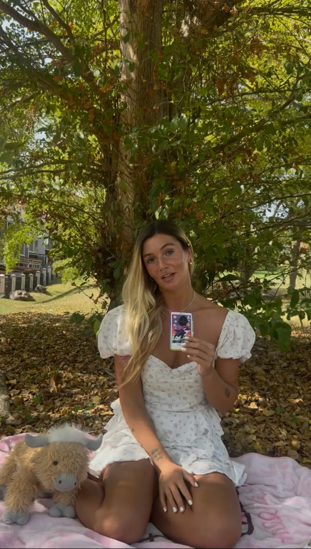 Gabriella sitting on a blanket beneath a tree, holding up a card and talking to the camera